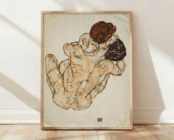 May include: Framed reproduction of an early 20th century expressionist drawing showing two embracing nude figures rendered in earthy tones of brown, beige and cream on textured paper background. The artwork features loose, sketchy linework and is displayed in a light wood frame against white walls on wooden flooring with natural window light casting diagonal shadows across the scene.