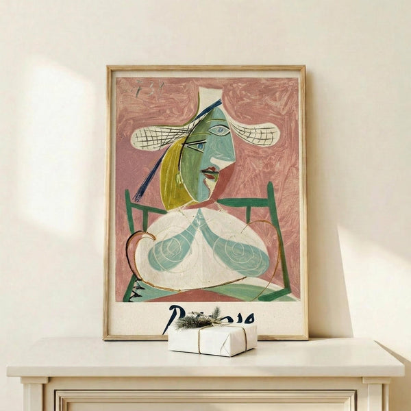 May include: Framed Picasso-style cubist portrait print displayed on light wood shelf against cream wall. Artwork features abstract female figure with green and yellow tones, geometric shapes, and pink background. Thin wood frame surrounds the poster. Small white wrapped gift box with natural twine and pine sprig sits on shelf beside the frame.
