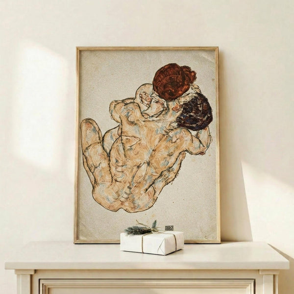May include: A framed vintage art print showing two embracing nude figures in earthy tones of beige, brown and reddish hues, displayed on a light wood surface next to a small white wrapped gift with greenery. The artwork features expressive line work in an early 20th century style, housed in a thin gold frame against a cream colored wall.