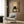 May include: Framed poster print featuring a black cat with wide white eyes sitting behind a bowl of ramen noodles, surrounded by vintage Japanese advertisements and bottles. The artwork is displayed on a wood-paneled wall above a cream-colored modern armchair with geometric throw pillow. A gold floor lamp and side table with open book complete the contemporary living room setting with neutral tones and natural lighting from nearby window.