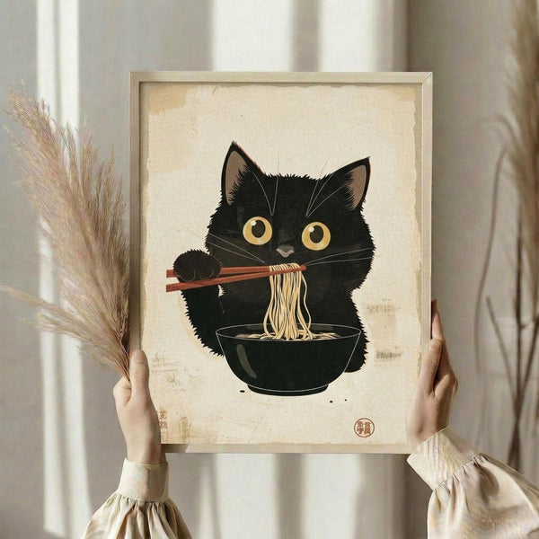 May include: Person holding framed art print featuring adorable black cat with golden eyes eating ramen noodles with chopsticks from dark bowl, illustrated in traditional Japanese ukiyo-e woodblock print style on cream aged paper background with red seal stamp, displayed in light wooden frame against neutral wall with dried pampas grass visible on left side