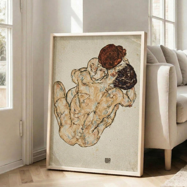 May include: A framed reproduction of an early 20th century expressionist drawing showing two embracing figures with auburn and dark hair rendered in earthy tones of beige, brown and muted blues. The artwork is displayed in a light wooden frame leaning against a white wall next to a beige upholstered armchair in a bright, naturally-lit room with large windows.