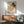 May include: Framed art print featuring two embracing figures in warm earth tones and muted colors, displayed above a mid-century modern wooden credenza with sliding doors. A white ceramic table lamp sits on the credenza, and a sage green upholstered armchair with wooden frame is positioned nearby. Natural sunlight casts geometric shadows across the white wall, creating a serene living room setting with vintage modern furniture.