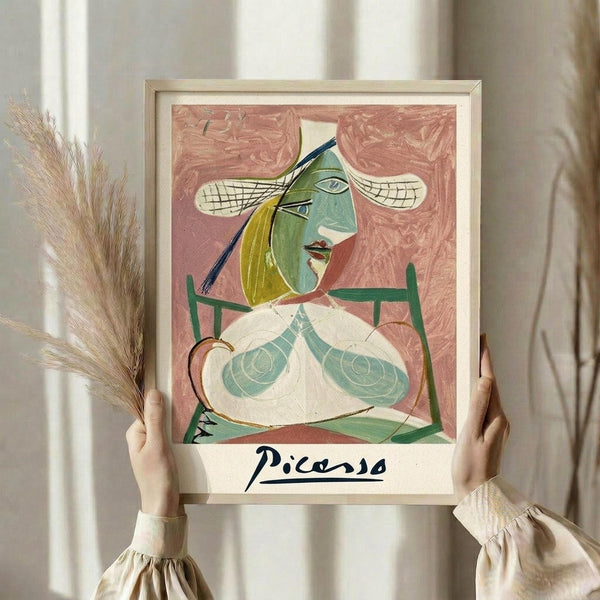May include: Person in cream clothing holding framed art print showing abstract portrait in Picasso's cubist style. Figure has green and yellow geometric face, bare torso, against pink background. Light wood frame with "Picasso" text below artwork. Dried pampas grass visible on left side, neutral curtains in background.