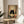 May include: Framed poster print displayed on light wooden round table next to beige upholstered chair and plant. Artwork shows black cat with large white eyes eating ramen noodles from ceramic bowl, surrounded by vintage Japanese restaurant advertisements and signage in muted colors. Natural sunlight streams across white wall creating soft shadows.