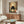 May include: Framed poster artwork featuring a black cat with large white eyes sitting behind a bowl of ramen noodles, surrounded by vintage Japanese text and food imagery in warm earth tones. The artwork is displayed above a mid-century modern walnut credenza in a minimalist living room with cream walls, white upholstered chair, brass floor lamp, and glass-top coffee table.