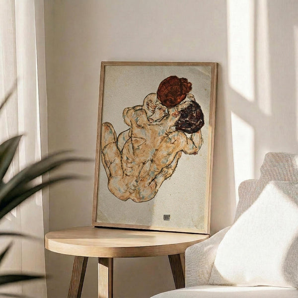 May include: Framed art print displaying an expressionist drawing of two embracing figures in earth tones and muted colors, positioned on a round wooden side table next to a beige upholstered chair. Natural sunlight streams across the neutral-toned room, casting soft shadows. A plant's leaves are visible in the foreground, creating an intimate living space setting.