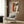 May include: Framed Picasso-style cubist poster with geometric face in orange, beige, and brown tones hanging on wood-paneled wall above cream upholstered armchair with geometric pillow. Modern floor lamp with brass finish and circular base stands nearby next to dark round side table with open book. Warm neutral interior with curtained window.