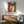 May include: Framed Picasso-inspired cubist face poster with geometric shapes in orange, brown, and cream tones hanging above a wooden sideboard. Modern living room setting with sage green armchair, white table lamp, and natural sunlight streaming across white walls creating dramatic shadows.
