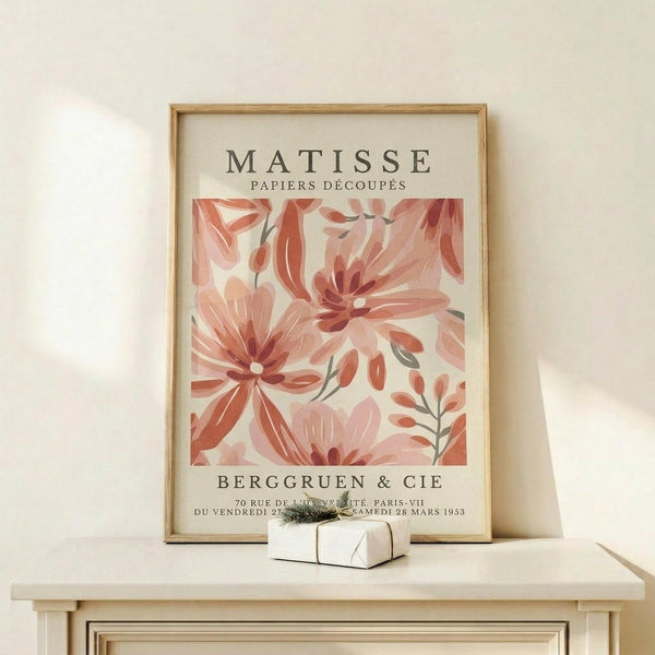 May include: Framed vintage-style art print featuring coral and pink cut-out flowers in Matisse's paper collage style, displayed on light wood shelf against cream wall. Small wrapped gift box with greenery sits beside the wooden-framed poster. Botanical design shows layered petals and leaves in warm peachy tones with exhibition text below the floral artwork.