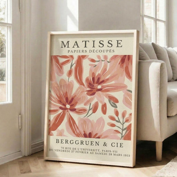 May include: Large framed poster leaning against white wall next to beige sofa in bright room with windows. Print features coral and pink abstract floral cutout shapes in Matisse's papier découpé style with cream background. French exhibition text at top and bottom in black letters. Light wooden frame, approximately 2-3 feet tall, positioned on light wood floors with natural sunlight streaming across scene.