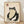 May include: Framed artwork leaning against white wall on light wooden floor. Print shows minimalist black ink illustration of rotund frog character sitting cross-legged with small bowl, rendered in traditional Japanese woodblock style on cream textured paper. Red square seal stamp in upper left corner. Natural wood frame with thin profile. Soft natural lighting creates subtle shadows on wall.