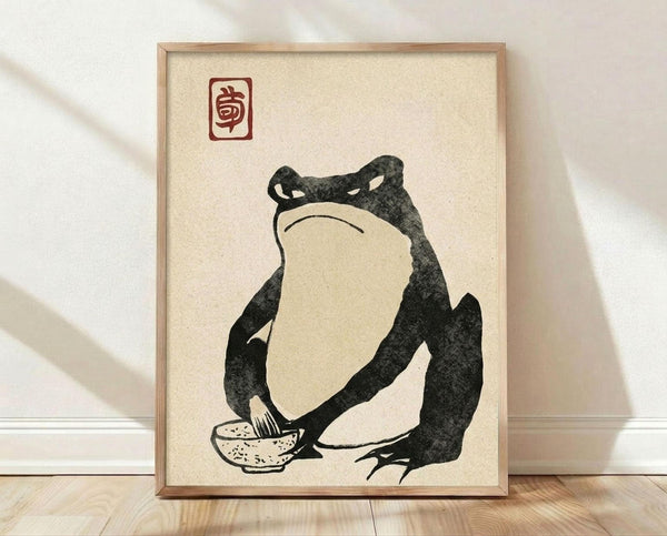 May include: Framed artwork leaning against white wall on light wooden floor. Print shows minimalist black ink illustration of rotund frog character sitting cross-legged with small bowl, rendered in traditional Japanese woodblock style on cream textured paper. Red square seal stamp in upper left corner. Natural wood frame with thin profile. Soft natural lighting creates subtle shadows on wall.