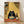 May include: Framed art print leaning against white wall on light wood floor. Shows black cat with large white eyes eating ramen noodles with chopsticks at orange table. Japanese-style setting with bamboo mat background, sake bottle, small glass, shelving with bottles. Warm yellow lighting, cartoon illustration style in thin wooden frame.