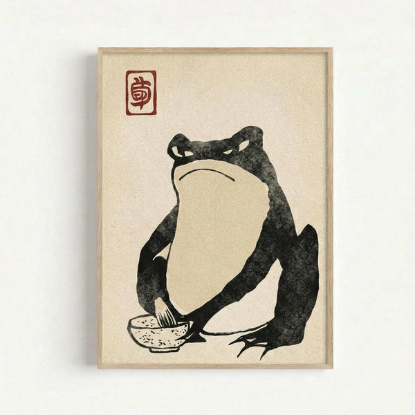May include: Framed Japanese-style artwork showing a large dark frog sitting with a small spotted bowl, rendered in black ink on cream textured paper. The minimalist illustration features bold brushstrokes and a red seal stamp in the upper left corner. The piece is displayed in a light wood frame against a white wall, casting a subtle shadow.