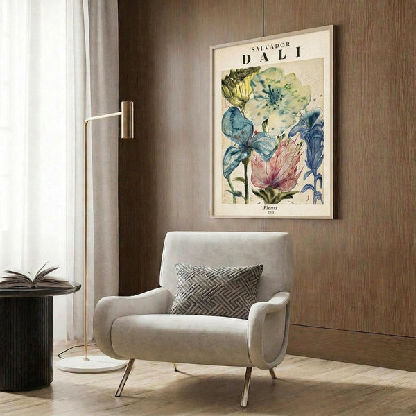 May include: A framed botanical poster featuring colorful vintage-style flowers in blues, greens, yellows, and pink with "Salvador Dali" text at top, displayed on wood-paneled wall above a beige upholstered armchair with geometric patterned pillow, brass floor lamp, and dark round side table with open book in a modern neutral-toned room setting.