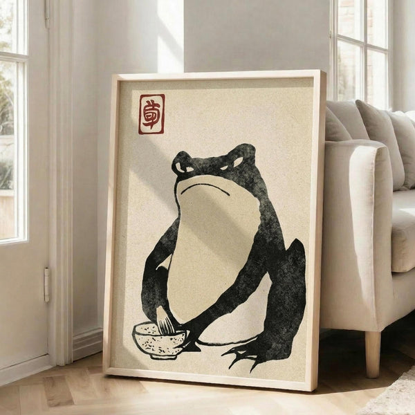 May include: Framed artwork displaying a stylized black frog with white belly sitting cross-legged, appearing grumpy with downturned mouth, holding what appears to be a small bowl. Red Japanese-style seal stamp in upper corner. Cream-colored background with natural wood frame, positioned against white walls near beige upholstered chair and large windows with natural lighting casting shadows across wooden floor.