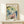 May include: Framed botanical art print featuring watercolor flowers in blue, green, yellow and pink tones with "Salvador Dali" text at top, displayed on light wood shelf against white wall with small white decorative object nearby. Vintage-style floral illustration with delicate petals and stems in soft, dreamy colors within natural wood frame.