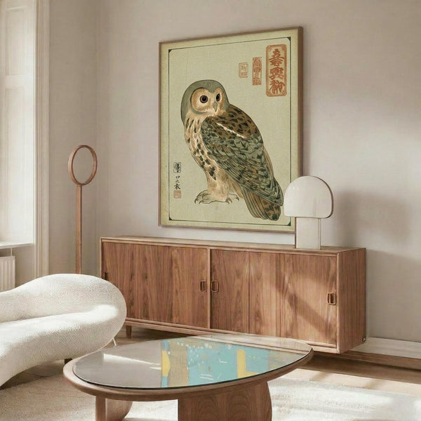 May include: Japanese woodblock-style owl print with traditional calligraphy and red seals, displayed in thin wooden frame above mid-century walnut credenza. Room features cream walls, white upholstered chair, oval glass coffee table with colorful abstract art underneath, and warm natural lighting creating soft shadows.