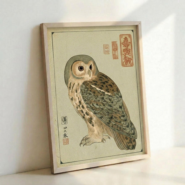 Framed Japanese woodblock-style illustration of an owl with cream and tan feathers, olive green wings, and distinctive round facial disc. The artwork features traditional red seal stamps with Japanese characters on pale green paper with a thin black border. The simple wooden frame leans against a white wall, casting soft shadows. The owl is depicted in a realistic pose showing detailed feather patterns and textures in muted earth tones.