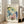 May include: Large framed botanical art print leaning against white wall beside beige upholstered chair. Artwork features watercolor-style flowers in blue, yellow, pink and green tones with "SALVADOR DALI" text at top and "Fleurs 1948" at bottom. Natural wood frame, cream matting. Bright room with white trim and hardwood floors.