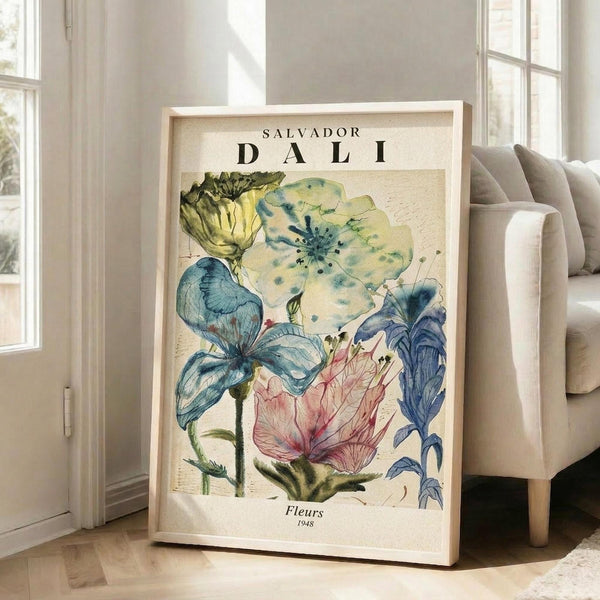 May include: Large framed botanical art print leaning against white wall beside beige upholstered chair. Artwork features watercolor-style flowers in blue, yellow, pink and green tones with "SALVADOR DALI" text at top and "Fleurs 1948" at bottom. Natural wood frame, cream matting. Bright room with white trim and hardwood floors.