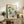 May include: Framed botanical artwork featuring colorful watercolor flowers in blues, greens, yellows and pink displayed on rustic wooden sideboard in neutral living room setting. Natural light illuminates cream walls, woven chair, potted fern, ceramic vases and stacked books creating warm, artistic home decor scene.