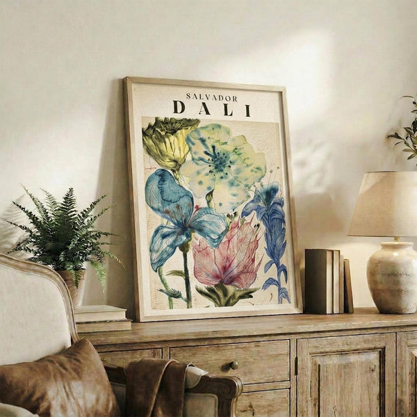 May include: Framed botanical artwork featuring colorful watercolor flowers in blues, greens, yellows and pink displayed on rustic wooden sideboard in neutral living room setting. Natural light illuminates cream walls, woven chair, potted fern, ceramic vases and stacked books creating warm, artistic home decor scene.
