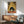 May include: Framed poster showing a black cat with wide white eyes eating ramen noodles from a bowl, set in a Japanese kitchen with bamboo mat background and sake bottle. Wooden credenza below holds white ceramic table lamp. Mid-century modern sage green armchair with wooden frame sits beside round coffee table. Bright natural light streams across white wall creating geometric shadows.