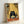 May include: Framed poster print leaning against white wall showing black cat with large white eyes eating ramen noodles with chopsticks. Cat sits at orange table with bowl of noodles, glass, and bottle labeled "SOJO". Background features yellow bamboo mat wall with small shelves holding bottles and containers. Bottom text reads "RELYSTES FULL ENERGY". Light wood frame, illustration style artwork in warm yellow and orange tones.