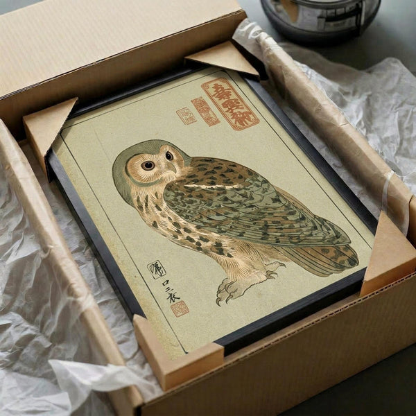 A vintage-style Japanese woodblock print of an owl with green and brown plumage and distinctive facial disc, featuring traditional red calligraphy seals. The artwork is displayed in a dark frame, carefully packaged in an open cardboard box with protective tissue paper and wooden dowels for shipping.