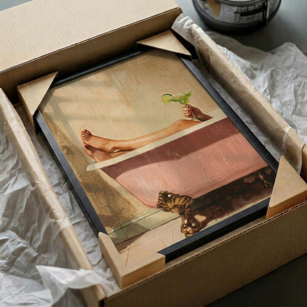 May include: Framed vintage-style poster artwork showing a person relaxing in a pink bathtub holding a green cocktail, with a small tiger nearby on tiled floor. The print has warm peachy-pink and beige tones with a dreamy, surreal quality. Black frame sits in cardboard shipping box with white tissue paper and bubble wrap packaging materials visible around it.