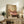 May include: Framed artwork showing a person's legs relaxing in a pink clawfoot bathtub while holding a green margarita cocktail, displayed on a rustic wooden sideboard alongside books, ceramic vases, and a fern plant in a warm, sunlit living room with neutral walls and cozy brown leather seating.