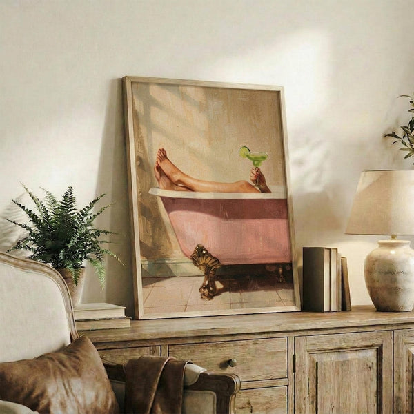 May include: Framed artwork showing a person's legs relaxing in a pink clawfoot bathtub while holding a green margarita cocktail, displayed on a rustic wooden sideboard alongside books, ceramic vases, and a fern plant in a warm, sunlit living room with neutral walls and cozy brown leather seating.
