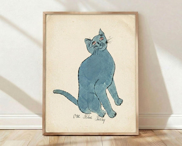 May include: Framed watercolor illustration of a blue cat sitting upright and looking up, painted in soft blue-gray tones with pink inner ears and eyes. The cat appears plump and content with whiskers and simple line details. Handwritten text below reads "One Blue Pussy" in cursive script. The artwork is displayed in a light wood frame against cream textured paper, leaning against a white wall on light wooden flooring with soft natural lighting casting shadows.
