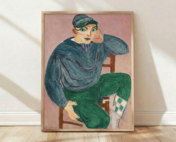 May include: Framed art print of a seated figure in teal blue top and green pants, wearing a cap, with hand positioned near face in contemplative pose. The artwork features bold brushstrokes and vibrant colors against a pink background. The print is displayed in a light wood frame, leaning against a white wall on light wooden flooring with soft natural lighting creating gentle shadows.