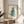 May include: Framed watercolor illustration of a blue cat sitting upright and looking up, displayed on a round wooden side table next to a beige upholstered chair. Natural sunlight streams across the neutral-toned room, casting soft shadows on the white walls. Green plant leaves visible in the corner, creating a cozy modern living space setting.