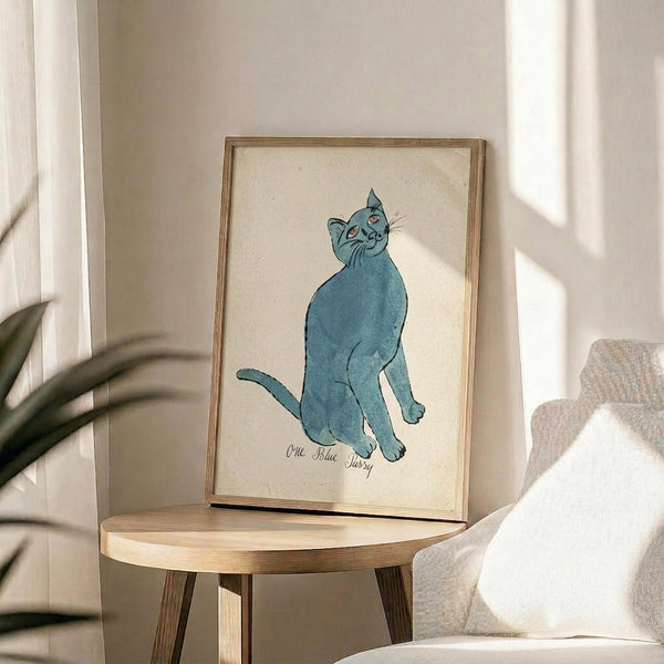May include: Framed watercolor illustration of a blue cat sitting upright and looking up, displayed on a round wooden side table next to a beige upholstered chair. Natural sunlight streams across the neutral-toned room, casting soft shadows on the white walls. Green plant leaves visible in the corner, creating a cozy modern living space setting.