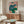 May include: Colorful portrait painting of seated figure in blue top and green pants on pink background, displayed above wooden credenza in modern living room with white curved chair, glass coffee table, and floor lamp