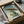 May include: Framed Japanese woodblock print artwork featuring a large green frog holding a bamboo umbrella in the rain, displayed in black frame inside cardboard shipping box with protective wrapping materials and wooden dowels, photographed on white fabric surface next to dark ceramic bowl