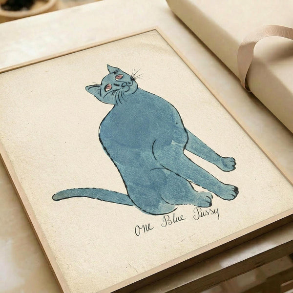 A whimsical watercolor illustration of a plump blue cat sitting upright with its head tilted back and eyes closed in a content expression. The cat is rendered in soft blue-gray tones with delicate line work defining its features, whiskers, and body. Below the cat is handwritten text reading "One Blue Pussy" in cursive script. The artwork is displayed on cream-colored paper within a simple wooden frame, photographed on a light wooden surface next to a roll of kraft paper and white ribbon.