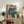 May include: Framed poster of a seated figure in teal sweater and green pants against pink background, displayed on rustic wooden sideboard alongside fern plant, ceramic vase, and books in neutral-toned living room setting