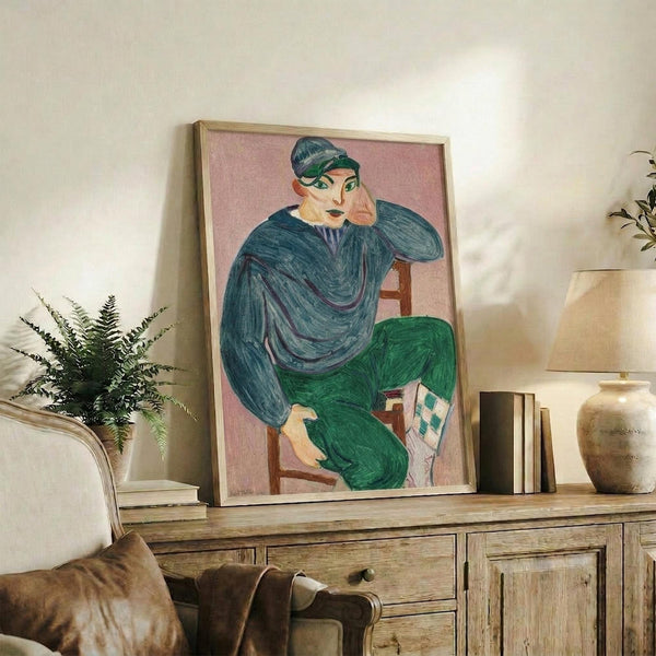 May include: Framed poster of a seated figure in teal sweater and green pants against pink background, displayed on rustic wooden sideboard alongside fern plant, ceramic vase, and books in neutral-toned living room setting