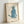 May include: Light wood frame displaying vintage-style illustration of blue cat sitting upright with head tilted back, rendered in soft blue watercolor with pink inner ears and whiskers, text below reads "One Blue Pussy" in cursive script, cream textured background, frame leaning against white wall