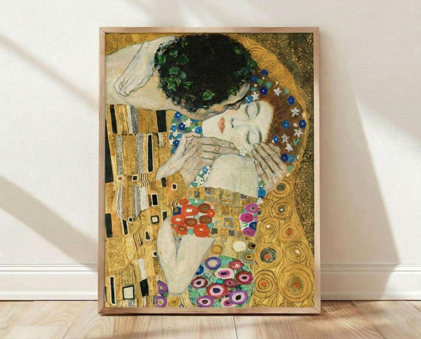 May include: Framed reproduction of Klimt's famous golden painting showing an embracing couple against ornate patterned background with geometric shapes and colorful circles, displayed in light wood frame leaning against white wall on wooden floor.