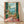 May include: Framed vintage-style Mediterranean balcony scene artwork leaning against white wall on light wood floor. Painting shows wicker chair on coral-pink tiled terrace with green shutters, tropical plants, and ocean view with distant mountains. Warm peachy-coral interior walls, decorative pottery, and palm fronds visible. Natural wood frame, soft window light creating shadows on wall.
