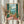 May include: Person holding framed vintage-style coastal artwork showing Mediterranean balcony scene with green shutters, wicker chair, potted pink flowers, palm fronds, and blue ocean view. Warm peachy-coral walls, decorative railing, distant mountains. Person wearing cream ruffled sleeves holds dried pampas grass, white curtained background.