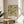 May include: Framed poster with ornate floral pattern featuring green foliage, red berries, yellow flowers, and decorative vines in vintage botanical style. Print displays on round wooden side table next to beige upholstered chair with white throw pillow. Natural sunlight streams across neutral-toned room with white curtains and potted plant visible in corner.