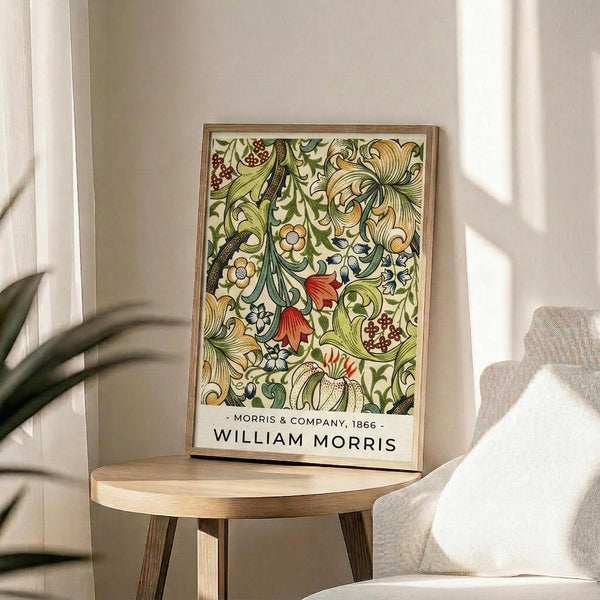 May include: Framed poster with ornate floral pattern featuring green foliage, red berries, yellow flowers, and decorative vines in vintage botanical style. Print displays on round wooden side table next to beige upholstered chair with white throw pillow. Natural sunlight streams across neutral-toned room with white curtains and potted plant visible in corner.