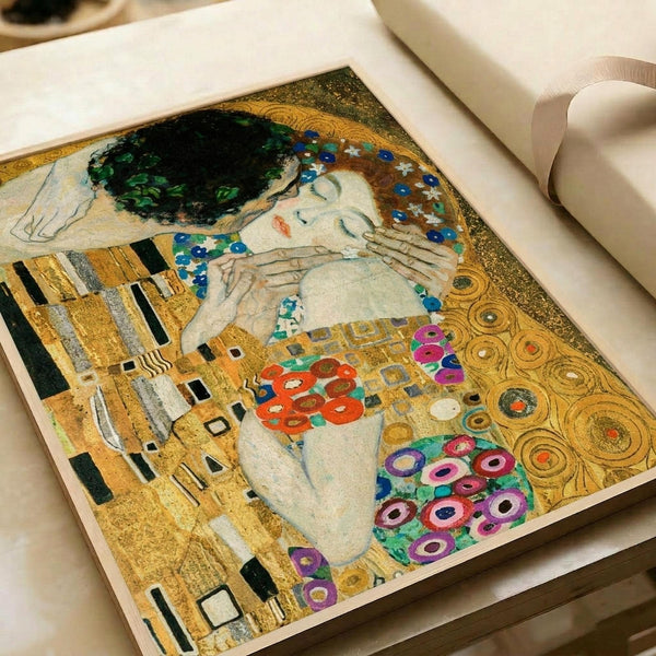 Reproduction of Gustav Klimt's famous painting "The Kiss" shown as a flat art print in an open book or portfolio. The artwork features two embracing figures against a golden background with ornate geometric patterns, swirling circles, and vibrant flowers in purple, red, and pink. The male figure wears gold robes with black rectangular patterns while the female figure is adorned with circular motifs and floral designs. Rich gold leaf effect dominates the composition with Art Nouveau styling cha